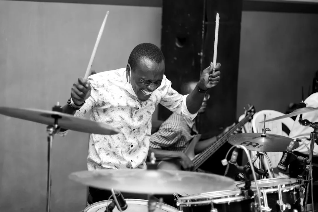 A drummer sitting behind a well-worn acoustic kit in a warmly lit rehearsal room, sticks in hand, surrounded by cymbals and a laptop open to a gear guide